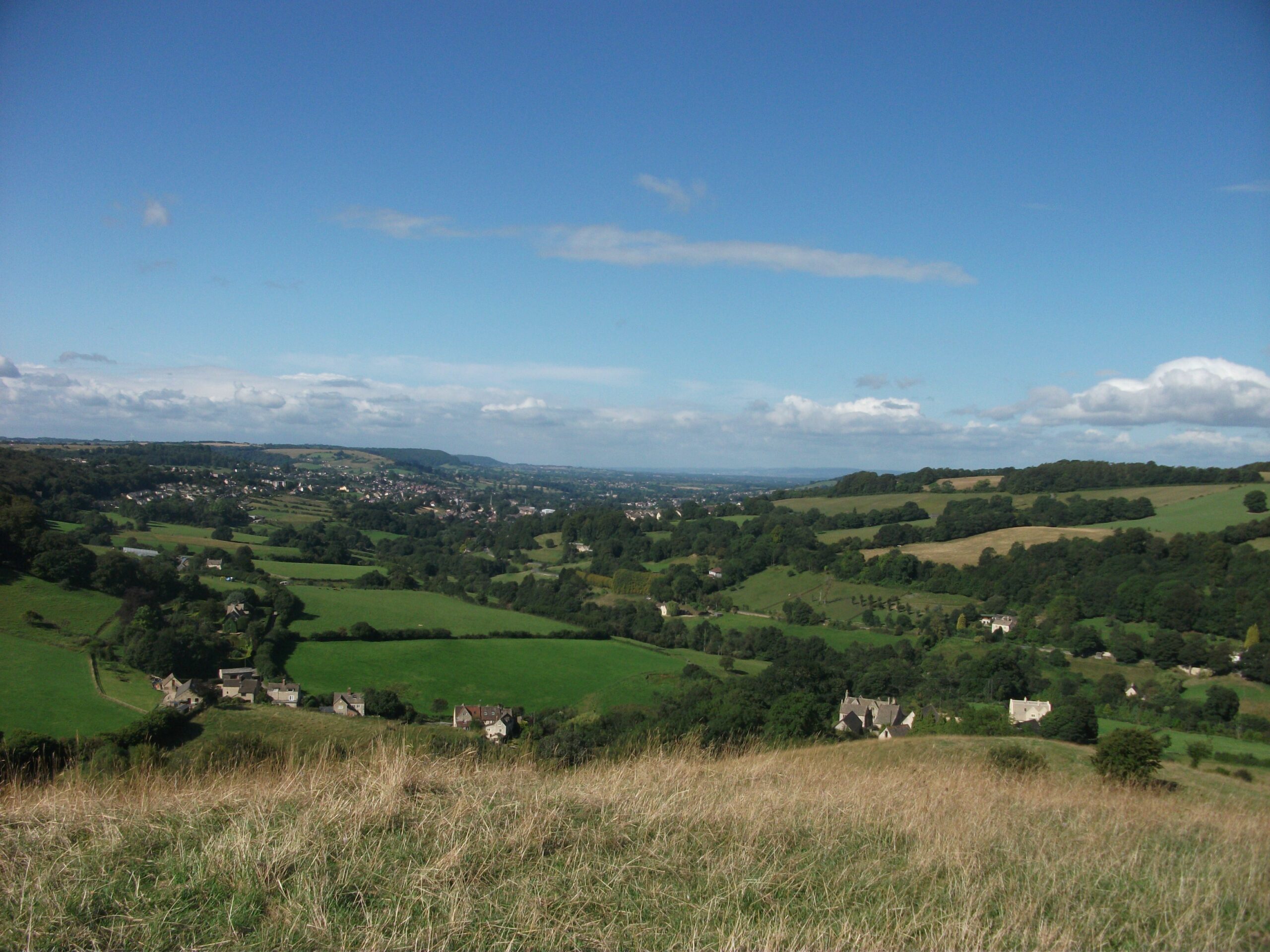 Looking down from the grassy top of Swift's Hill over the green Slad valley, with scattered houses and woods under a blue sky
