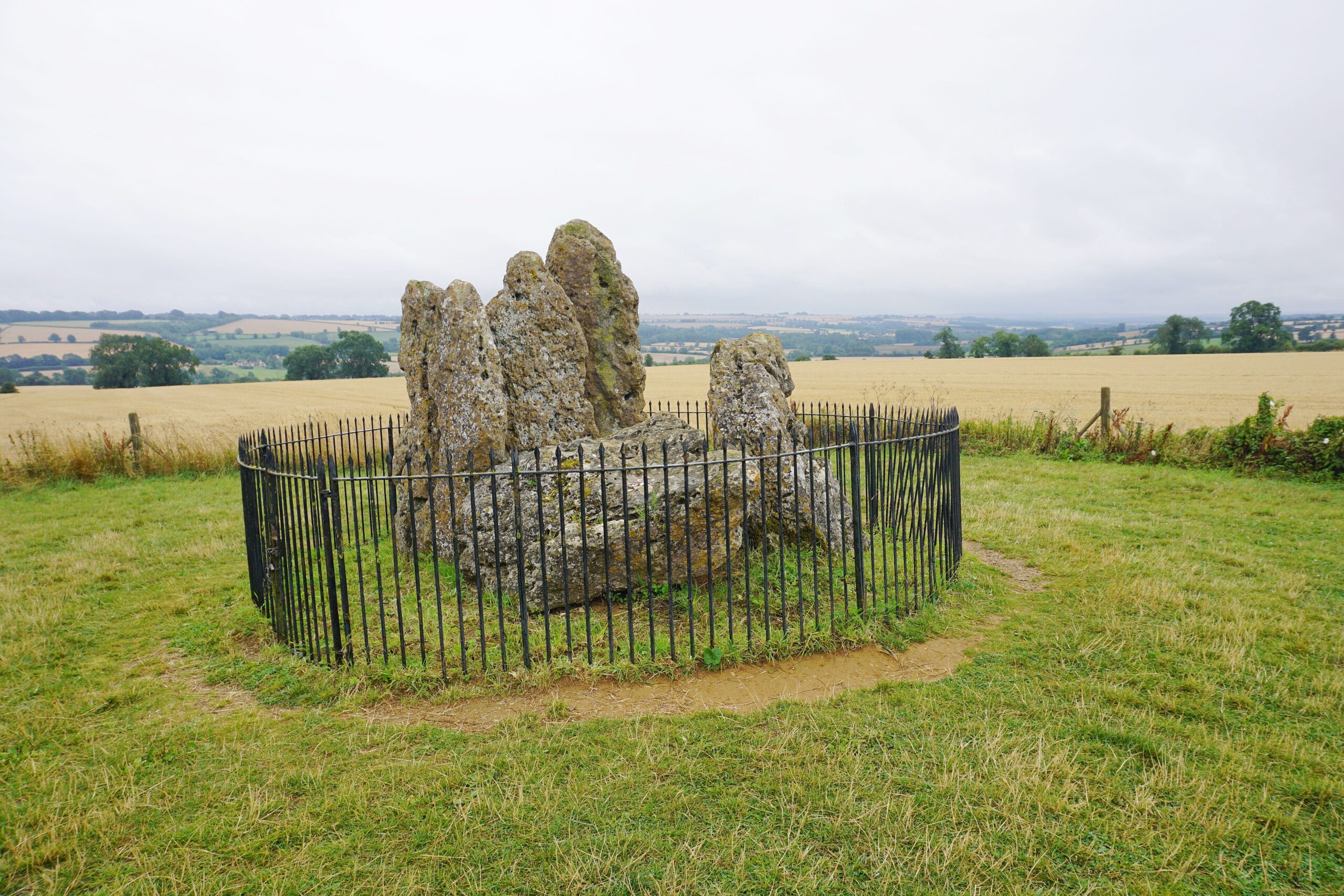 The Whispering Knights, a cluster of tall weathered standing stones inside iron railings, with farmland stretching away behind